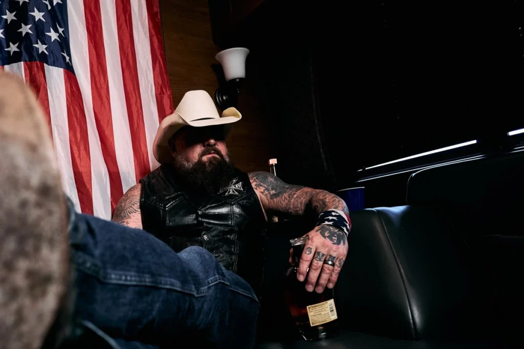 Creed Fisher in a white cowboy hat and leather vest, seated in front of an American flag