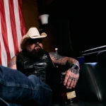 Creed Fisher in a white cowboy hat and leather vest, seated in front of an American flag