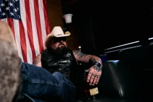 Creed Fisher in a white cowboy hat and leather vest, seated in front of an American flag